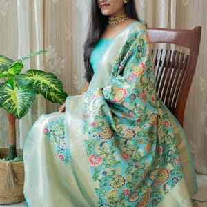 May include: A light blue silk saree with a floral pattern in shades of pink, yellow, green, and blue. The saree has a silver border and is draped over a chair.