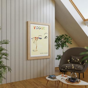 May include: A framed "VOGUE" poster with a fashion illustration of an eye and hand applying makeup. The poster is in a light wood frame and hangs on a light gray paneled wall. The room has a modern design with a brown chair and small tables.