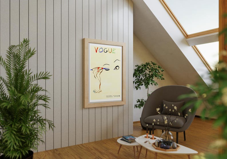 May include: A framed "VOGUE" poster with a fashion illustration of an eye and hand applying makeup. The poster is in a light wood frame and hangs on a light gray paneled wall. The room has a modern design with a brown chair and small tables.