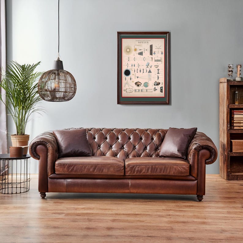 May include: A brown leather chesterfield sofa with tufted buttons and rolled arms sits in a living room with a framed vintage illustration of optical illusions and a hanging wicker pendant light.