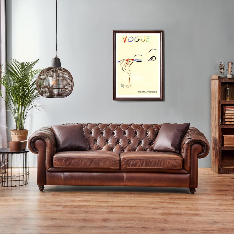 May include: A brown leather Chesterfield sofa with two matching throw pillows. A framed "Vogue" magazine cover art print hangs above the sofa. A decorative cage-style pendant light hangs from the ceiling. A potted plant sits to the left.