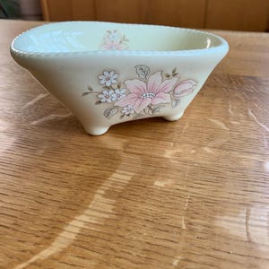 May include: A cream-colored ceramic bathtub-shaped dish featuring a delicate floral pattern with pink and white flowers on the side. The vessel has a textured beaded rim, a smooth interior, and stands on four small, rounded feet for stability.