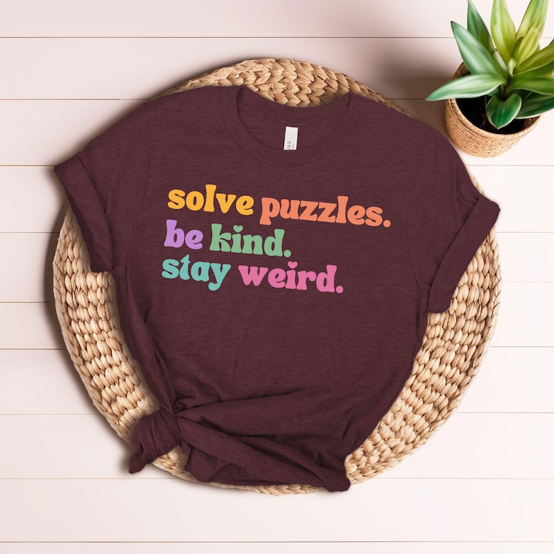 Puede incluir: Camiseta color burdeos con el texto "solve puzzles. be kind. stay weird." en colores arco&iacute;ris.