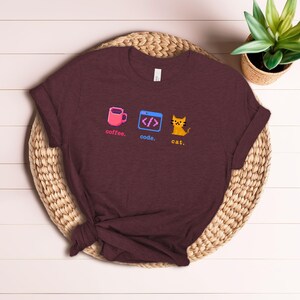 K&ouml;nnte beinhalten: Burgunderfarbenes T-Shirt mit pixeligen Grafiken: eine rosa Kaffeetasse, ein Code-Symbol und eine gelbe Katze. Die W&ouml;rter "coffee", "code" und "cat" sind darunter gedruckt. Das Shirt liegt auf einer gewebten Matte.