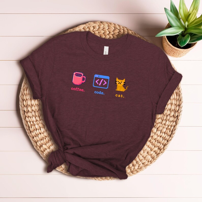 K&ouml;nnte beinhalten: Burgunderfarbenes T-Shirt mit pixeligen Grafiken: eine rosa Kaffeetasse, ein Code-Symbol und eine gelbe Katze. Die W&ouml;rter "coffee", "code" und "cat" sind darunter gedruckt. Das Shirt liegt auf einer gewebten Matte.