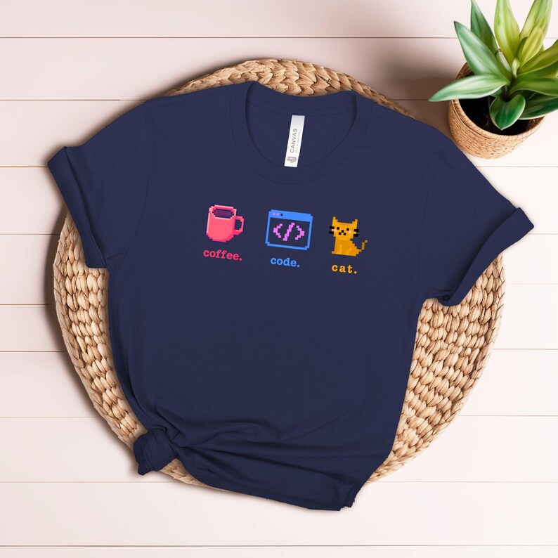 K&ouml;nnte beinhalten: Ein marineblaues T-Shirt mit einem pixeligen Design, das eine rosa Kaffeetasse, ein Codefenster und eine gelbe Katze zeigt. Die W&ouml;rter "coffee", "code" und "cat" sind in Rosa gedruckt.