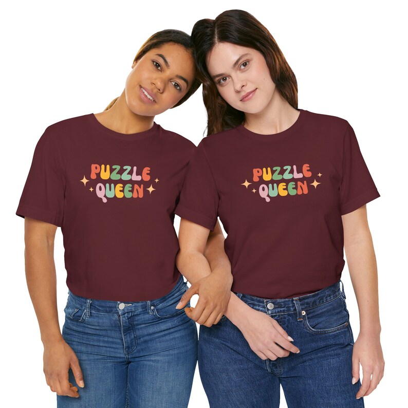 May include: Two people wearing maroon t-shirts with the text "Puzzle Queen" in colorful retro lettering. The shirts have a distressed look and are worn with blue jeans.
