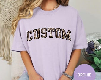 May include: A lavender t-shirt with the word "CUSTOM" printed in black letters with a leopard print pattern.