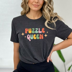 May include: A dark grey t-shirt with the text "PUZZLE QUEEN" in rainbow colors. The text is in a retro font style with stars on either side.