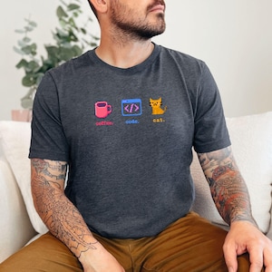 K&ouml;nnte beinhalten: Dunkelgraues T-Shirt mit einem pixeligen Design, das eine rosa Kaffeetasse, Code-Klammern und eine gelbe Katze zeigt. Die Worte "coffee. code. cat." sind darunter gedruckt. Das Shirt ist aus weichem Material.