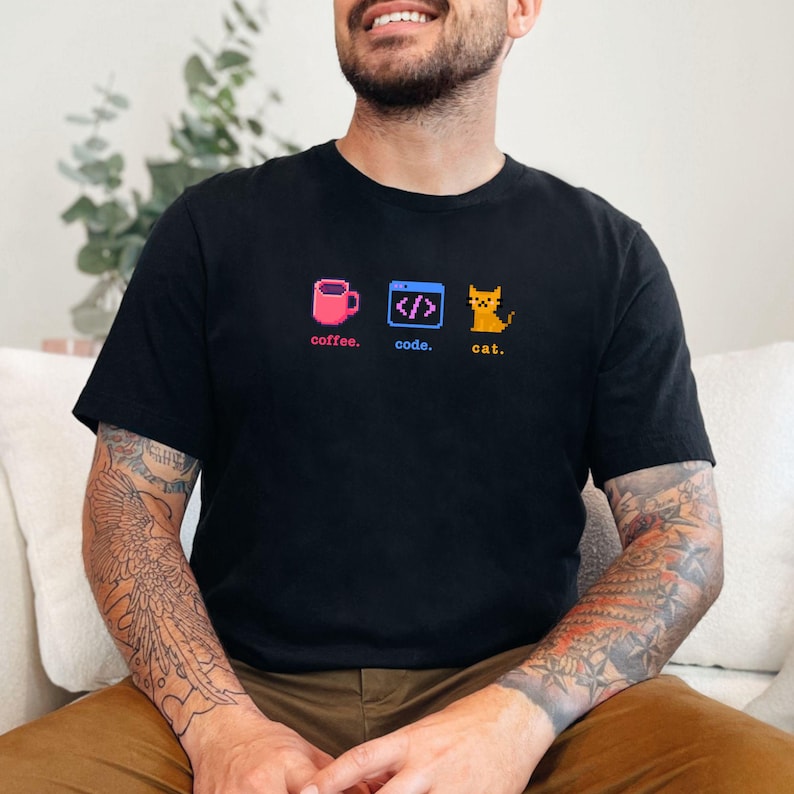 K&ouml;nnte beinhalten: Schwarzes T-Shirt mit einem pixeligen Design, das eine rosa Kaffeetasse, ein Code-Symbol und eine gelbe Katze zeigt. Die W&ouml;rter "coffee", "code" und "cat" stehen darunter. Das Shirt ist aus weichem Material.