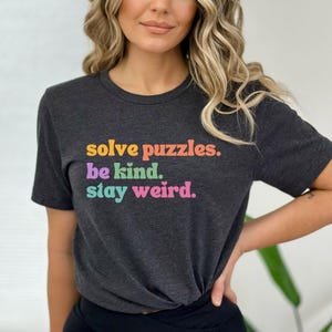 Puede incluir: Una camiseta gris oscuro con el texto "solve puzzles. be kind. stay weird." impreso en colores arco&iacute;ris.