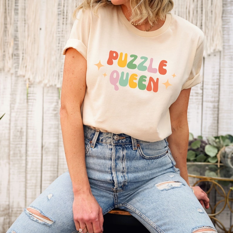 May include: A light beige t-shirt with the text "Puzzle Queen" in rainbow colors. The text is in a retro font style with a sparkle on each side.