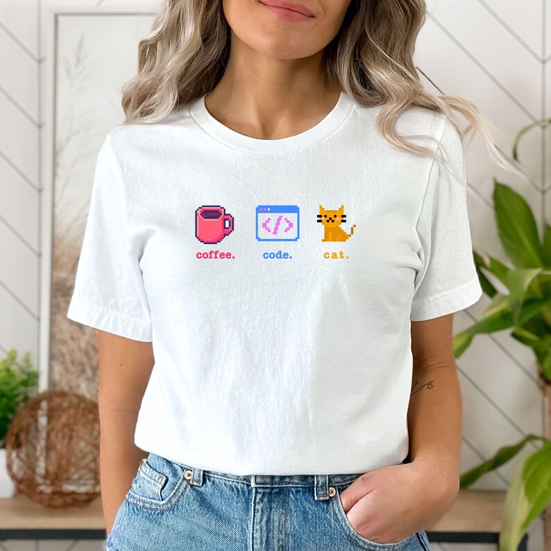 K&ouml;nnte beinhalten: Wei&szlig;es T-Shirt mit pixeligen Grafiken: eine rosa Kaffeetasse, ein Code-Symbol und eine orange Katze, mit den Worten "coffee. code. cat." darunter. Ein l&auml;ssiges Kleidungsst&uuml;ck f&uuml;r den Alltag.