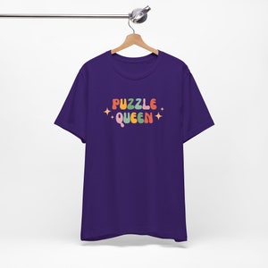 May include: Purple t-shirt with the text "PUZZLE QUEEN" in rainbow colors with stars on either side of the text.