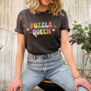 May include: A black t-shirt with the text "PUZZLE QUEEN" in rainbow colors. The text is in a retro font style with stars on either side.