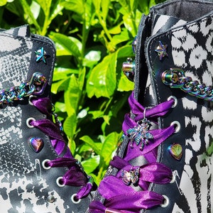 Puede incluir: Un par de botas de cuero negro con un diseño de estampado animal blanco y negro. Las botas tienen cordones de cinta morada con abalorios decorativos, incluyendo una abeja, una mariposa y un corazón. Las botas tienen un detalle de costura amarilla alrededor de la suela.