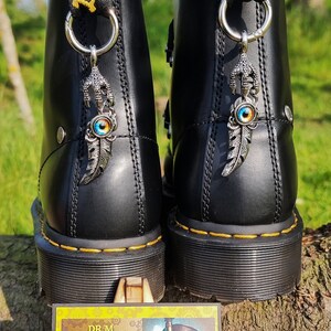 May include: A pair of black leather boots with yellow and black straps. Each boot has a silver charm with a blue eye and a feather attached to it. The boots are sitting on a wooden surface. The text on the business card reads "Dr M Retro Fitter".