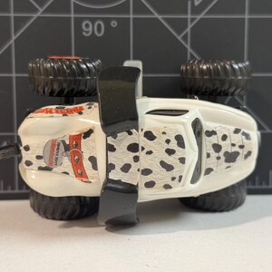 May include: A white and black toy monster truck with black wheels and a black spoiler. The truck has a dalmatian-like pattern on the sides and the text "Sports Rikt" on the side.