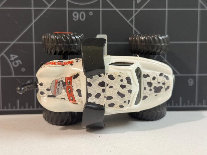 May include: A white and black toy monster truck with black wheels and a black spoiler. The truck has a dalmatian-like pattern on the sides and the text "Sports Rikt" on the side.