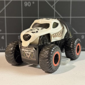 May include: A white and black toy monster truck with a dalmatian dog design. The truck has large black tires with orange rims and the words "Monster Jam" on the front.
