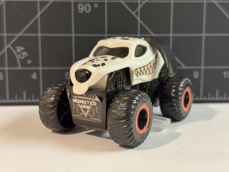 May include: A white and black toy monster truck with a dalmatian dog design. The truck has large black tires with orange rims and the words "Monster Jam" on the front.