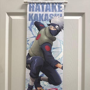 May include: A wall scroll featuring the character Hatake Kakashi from the anime series Naruto. The scroll displays a full-body illustration of the character in a dynamic pose, with the word "NARUTO" in orange at the bottom.