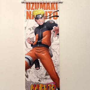 May include: A vertical wall scroll featuring a vibrant illustration of a character from the anime series Naruto. The character is depicted in an action pose, wearing an orange and black outfit. The scroll has the text "UZUMAKI NARUTO" at the top and "NARUTO" at the bottom.