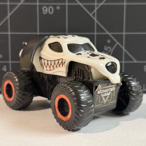 May include: A toy monster truck with a white and black dalmatian dog design. The truck has large black tires with orange rims and the words "Monster Jam" on the side.