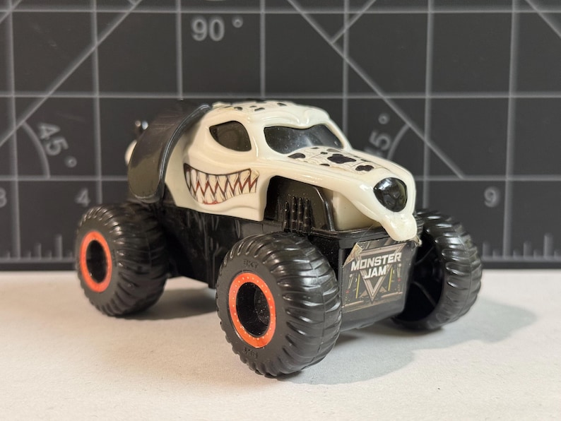 May include: A toy monster truck with a white and black dalmatian dog design. The truck has large black tires with orange rims and the words "Monster Jam" on the side.