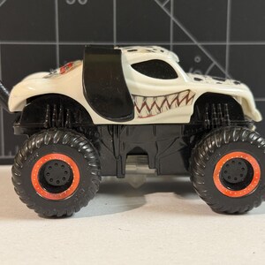 May include: A toy monster truck with a white body, black accents, and a menacing grin design. The truck has large black tires with orange rims. The vehicle is a collectible toy.