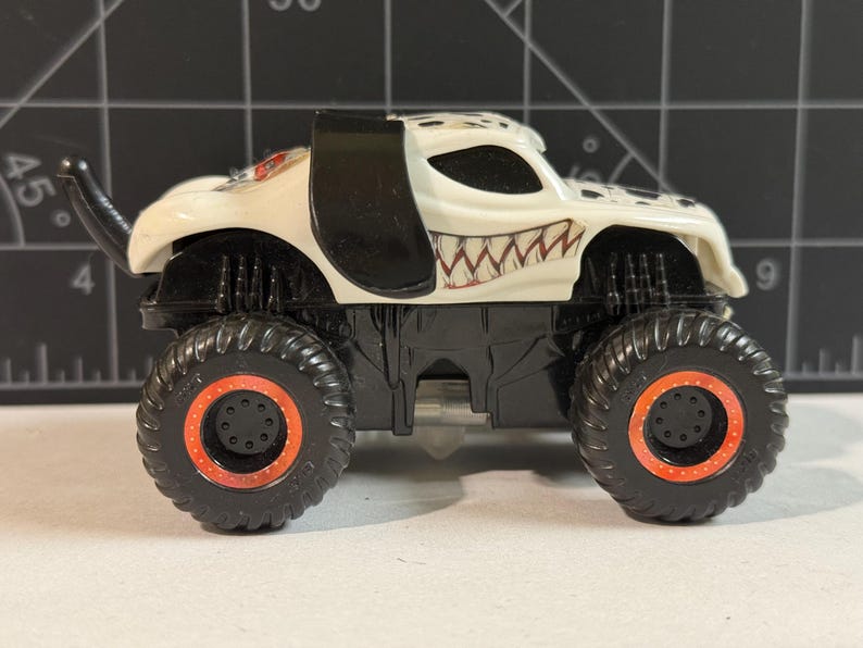 May include: A toy monster truck with a white body, black accents, and a menacing grin design. The truck has large black tires with orange rims. The vehicle is a collectible toy.