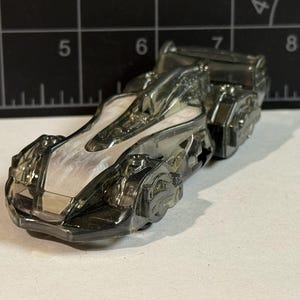 May include: A transparent, futuristic-looking toy race car. The car has a sleek design with a white interior and dark gray exterior. The toy car is on a white surface with a black ruler in the background.