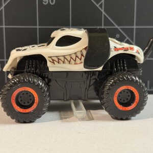 May include: A white and black monster truck toy with large black tires featuring orange rims. The truck has a dalmatian-like design with a toothy grin on the front. The text "Monster Jam" is visible on the side.