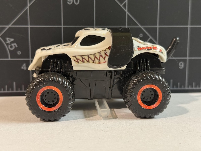 May include: A white and black monster truck toy with large black tires featuring orange rims. The truck has a dalmatian-like design with a toothy grin on the front. The text "Monster Jam" is visible on the side.