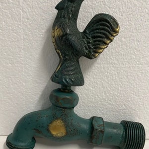 May include: A green metal garden faucet with a rooster design. The rooster is gold and green and has a raised wing. The faucet is made of metal and has a rustic look.