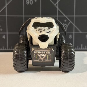 May include: A toy monster truck with a Dalmatian dog design. The truck has large black tires and a white body with black spots. The front of the truck features the "Monster Jam" logo.