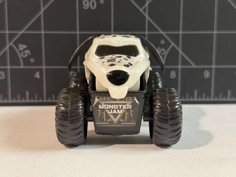 May include: A toy monster truck with a Dalmatian dog design. The truck has large black tires and a white body with black spots. The front of the truck features the "Monster Jam" logo.