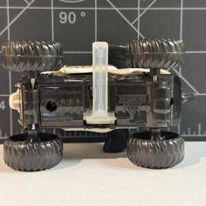 May include: A black toy monster truck with large, textured black tires. The underside of the truck is visible, showing the chassis and a clear plastic piece. The truck has a white and beige body.