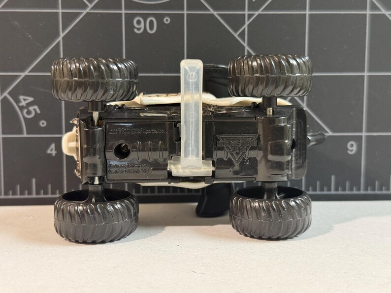 May include: A black toy monster truck with large, textured black tires. The underside of the truck is visible, showing the chassis and a clear plastic piece. The truck has a white and beige body.