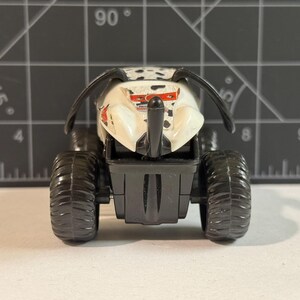 May include: A toy monster truck with a white body, black spots, and large black tires. The truck has a black front bumper and two curved black antennae. The toy is designed to look like a monster truck.