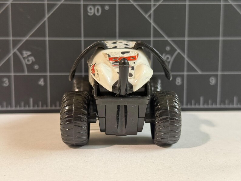 May include: A toy monster truck with a white body, black spots, and large black tires. The truck has a black front bumper and two curved black antennae. The toy is designed to look like a monster truck.