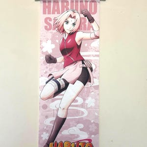 May include: A pink wall scroll featuring the anime character Haruno Sakura from Naruto. The character is depicted in a dynamic pose, wearing a red top, pink skirt, and black shorts. The scroll has the text "HARUNO SAKURA" and "NARUTO" in bold letters.