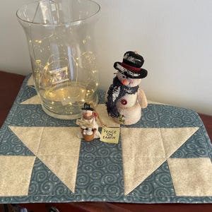 May include: A blue and cream quilted table runner with a starburst pattern. Two small snowman figurines are on the runner, one holding a sign that says "Peace on Earth".