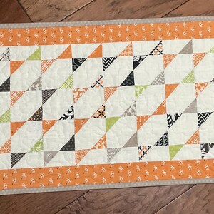 May include: A rectangular quilted table runner with a cream-colored center featuring a repeating pattern of orange, black, gray, and green triangles. The runner is bordered by a patterned orange fabric and a neutral binding.