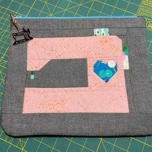 May include: A gray fabric zippered pouch with a pink sewing machine applique. The sewing machine has a blue heart detail. A small charm of a sewing machine hangs from the zipper. The pouch is on a green cutting mat.