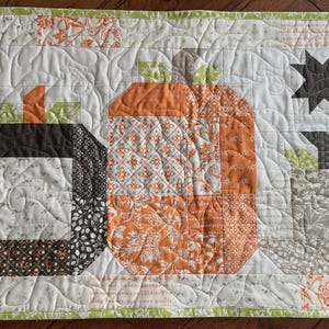 Puede incluir: Un camino de mesa acolchado con tres diseños de calabazas en tonos naranja, marrón y beige. Las calabazas están sobre un fondo blanco con un borde verde. También aparece una estrella oscura.