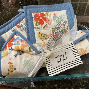 May include: An oversized hot pad set with a floral and chicken pattern, presented in a glass baking dish. The set includes two square hot pads and a triangular one, all with blue trim. A whisk and a tag that says "oversized hot pad" are included.