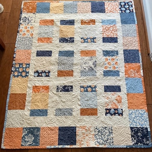 May include: A quilt with a patchwork design featuring squares of various floral patterns in shades of blue, orange, and cream. The quilt is quilted with a swirling pattern.