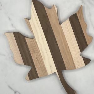 May include: A maple leaf shaped cutting board made of various wood types, including walnut, maple, and cherry. The board has a natural wood finish and is perfect for serving cheese, charcuterie, or other appetizers.